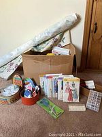Box with fabric rolls, vintage sewing patterns, circular cutter, tins of sewing notions, and other sewing accessories displayed on carpet.