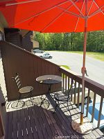 Outdoor patio set including two metal mesh chairs, round metal mesh table with central hole for umbrella, and large orange/red freestanding umbrella with stone base stand on wood deck.