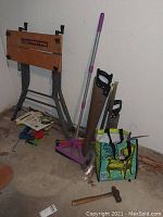 Full view showing workbench, saws, broom, reusable grocery bag with contents, and hammer on the floor.