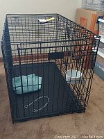 Black wire metal dog crate with front door, plastic tray, light blue cloth, white bowl, and chain collar inside, placed on carpeted floor.