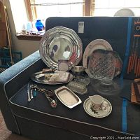 Full lot displayed on blue chair showing various metal and glass serving dishes, porcelain pieces, utensils and napkin holders
