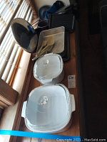 Two white Corning Ware covered dishes with floral design, waffle/sandwich press, metal baking pans, plastic utensils on wooden surface by window.