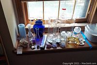 View of varied glassware collection including vases in three colors, pitcher, candle holders, candles, bottles and glass lidded containers arranged on a windowsill.