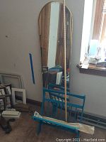 Full view of tall arched wall mirror leaning against the wall next to blue collapsible magazine rack and other items