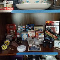 Several home improvement items on a shelf including various light bulbs, caulk, wall cushions, small containers, black binoculars, and a Timex clock.