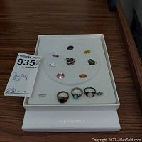 Top-down photo of the opened jewelry box showing three sterling silver rings, one pendant, and multiple interchangeable faux gemstones in various colors on a white insert.