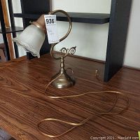 Desk lamp with metal curved base and ribbed glass shade on wooden surface next to shelving unit, showing the lamp profile and power cord.