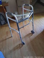 Foldable aluminum walker with two front wheels, viewed on wooden floor, showing padded hand grips and metal frame.