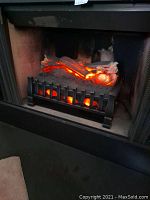 Electric log insert placed in fireplace opening, showing glowing ember light effect through logs and metal grille.