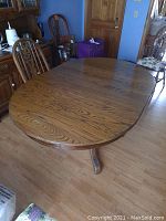 View of oval wood pedestal dining table with leaf inserted, showing rounded edges and wood grain pattern on tabletop surface.
