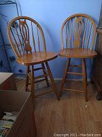 Two wooden bar stools with light-medium brown finish, spindle backs, two lower rungs each, swivel seats. One stool missing rear rung.