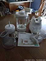 Cuisinart blender and food processor components arranged on table including pitcher, processor bowl, blades, lids, and instruction booklet