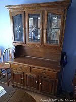 Front view of wood hutch showing upper leaded glass cabinet doors with decorative ovals, middle work surface, and lower drawers and cabinets. Shows brass hardware and oak finish.