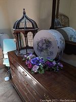 Wooden octagonal bird cage, round floral hat box, and assorted faux flowers displayed on a wooden dresser.