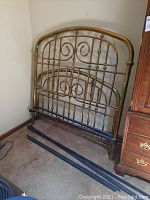 Brass headboard standing upright against wall with visible ornate swirl and rod details.