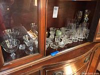 View of left side of cabinet showing stemmed wine glasses and some salt dips inside glass-front wooden cabinet