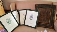 Full view of four framed wild flower prints and one framed carved wood relief on beige patterned sofa.