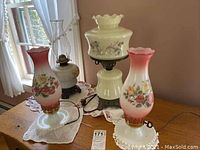 Photo shows three hurricane lamps, two with pink and white floral glass shades on brass bases, and one original oil lamp with clear chimney and dark metal base, placed on a wooden table with white doilies beneath them.
