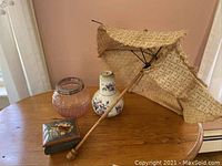 Photo showing antique beige parasol with fringe detail, clear round glass flower frog, white ceramic carafe and cup with purple floral decorations, and small painted wooden music box on round wooden table.