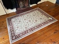 Full view of the rectangular polypropylene rug laid on a wooden floor showing beige center with floral patterns and red floral border.