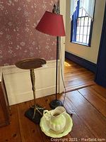Full view of brass chicken leg-shaped plant or cocktail stand, vintage floor lamp with red shade, and porcelain wash basin set on wooden floor.