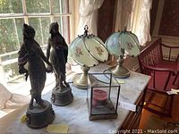 Photo of two vintage female figurines, two glass shade lamps with floral design, and a glass candle holder on marble table near window