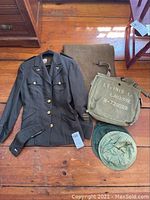 Full view of the WWII women's military jacket, side cap, blanket, olive canvas bag with stenciled name and number, and mosquito hat laid out on a wooden floor.