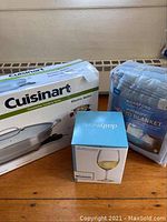 Photo displaying original boxed Cuisinart electric skillet, boxed Dailyware wine glasses, and packaged Guildford Technologies electric blanket on a hardwood floor.