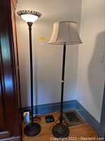 Two floor lamps standing against a wall in a room; left lamp has a frosted glass bowl shade with a decorative metal rim, right lamp has a cream fabric shade.