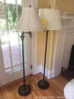 Two floor lamps standing side by side near a glass door and wall, showing full height and base, lampshades cream colored, metal base dark finished