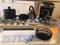 Set of multiple Swiss Diamond pots and pans with glass lids, kettle, and metal mesh strainers on kitchen countertop and shelf.