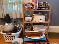 Photo of various kitchen items arranged on shelf and white plastic laundry basket showing CorningWare platters, roasting pan, crock pot, rice cooker, baking pans and metal kitchen utensils inside plastic basket.