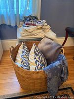 Photo showing assorted table linens stacked on a stool behind a large woven basket containing two blue and white patterned throw pillows and a gray blanket.