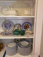 Overall view of shelves with assorted plates and serving pieces including white tureen, gravy boat, rectangular platter, colorful plates with floral patterns, and green sugar and creamer set