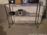 Front view of metal three shelf utility cart placed against a stone wall, showing empty wire shelves and metal legs on casters.