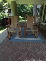 Two wooden rocking chairs with woven rattan seats and backs shown together on a porch rug. The chairs have spindle legs and finial-topped back posts. Weathered wood finish visible.