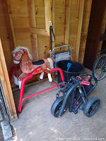 View of plush child’s riding horse on red metal frame rocking stand, folded black golf cart, aluminum frame folding chair, black bucket, and blue shovel in wooden shed.
