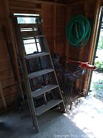 6 ft. wooden folding stepladder leaning against wall inside shed, accompanied by wrought iron style planter rack, green garden hose coiled on wall, and owl decoy statue on floor.