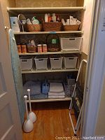 View of closet shelving containing toiletries in baskets on top shelf and middle shelf, white storage bins on middle shelf, towels and bath mats on bottom shelf, and two plunger-type items on the floor.