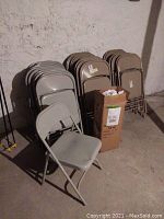 Three stacks of metal folding chairs in light gray/beige with a cardboard box of slipcovers in front of a basement wall