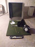 Full indoor golf practice setup showing mat with golf balls, driver, weighted bag, and foldable swing box frame.