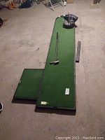 Long shot showing full indoor putting green mat with golf clubs and other accessories placed on it, on concrete floor.