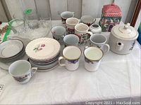 Photo showing assortment of china items including mugs, bowls, teapot on a white cloth background