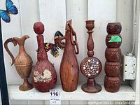 Photo showing five carved and inlaid artisan bottles and wooden decorative figures arranged side by side against white wall with butterfly decals.