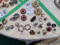 Photo showing assortment of novelty rings, mismatched earrings, and a jewelled floral pin on light fabric.
