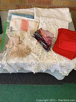 First photo showing a variety of linens including crocheted doilies, folded napkins, lace and plain tablecloths in white, cream, red, and multi-color.