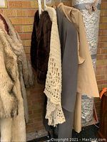 Photo showing side view of five hanging outerwear items against brick wall: beige faux fur jacket, dark brown fur jacket, beige crochet shawl, grey coat, and beige coat.