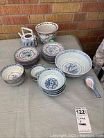 Full set of Asian style ceramic plates, bowls, spoon, planter, and teapot/creamer arranged on table against brick wall.