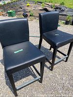 Front and side view of two black leather chairs with cushioned seats and backs and wooden legs placed outdoors on asphalt.