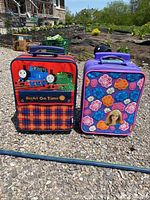 Two children's suitcases outdoors on gravel, one Thomas the Tank Engine with red plaid design and one Barbie floral purple suitcase with doll face on front.